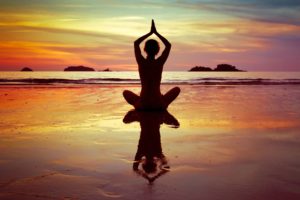 A woman on the beach at sunset practicing yoga, sitting in lotus pose.